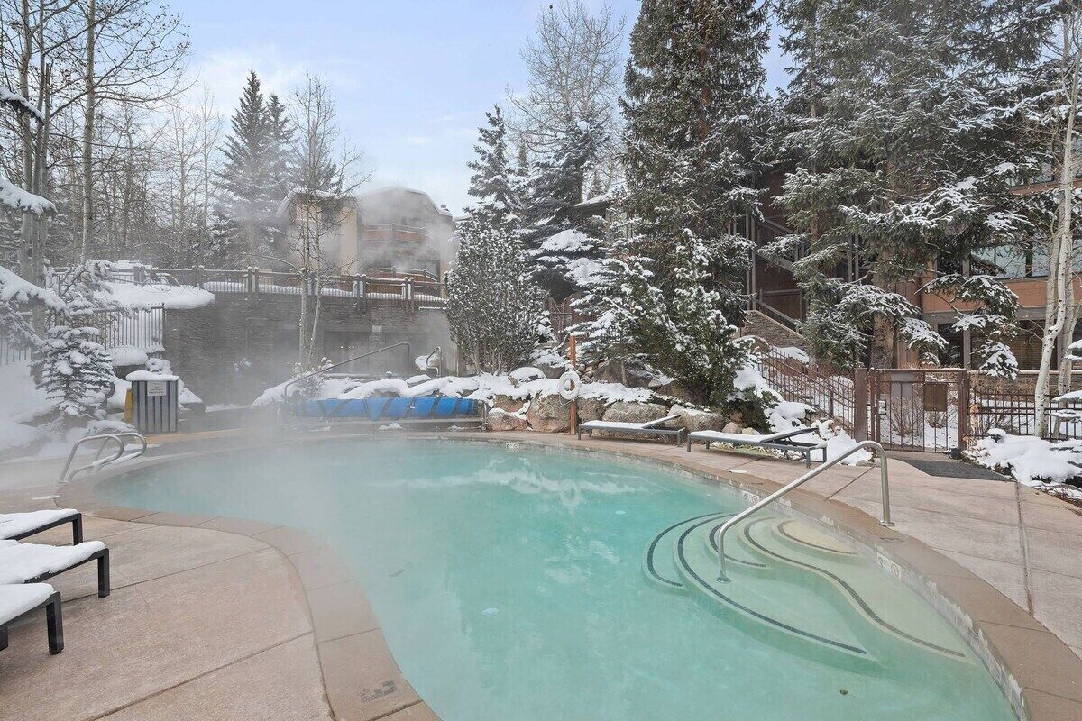 A hot tub surrounded by snow-covered trees and a building in the background.