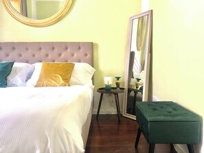 Un lit bien fait, doté d’une tête de lit capitonnée, une table de chevet avec une lampe, un miroir et un pouf vert.