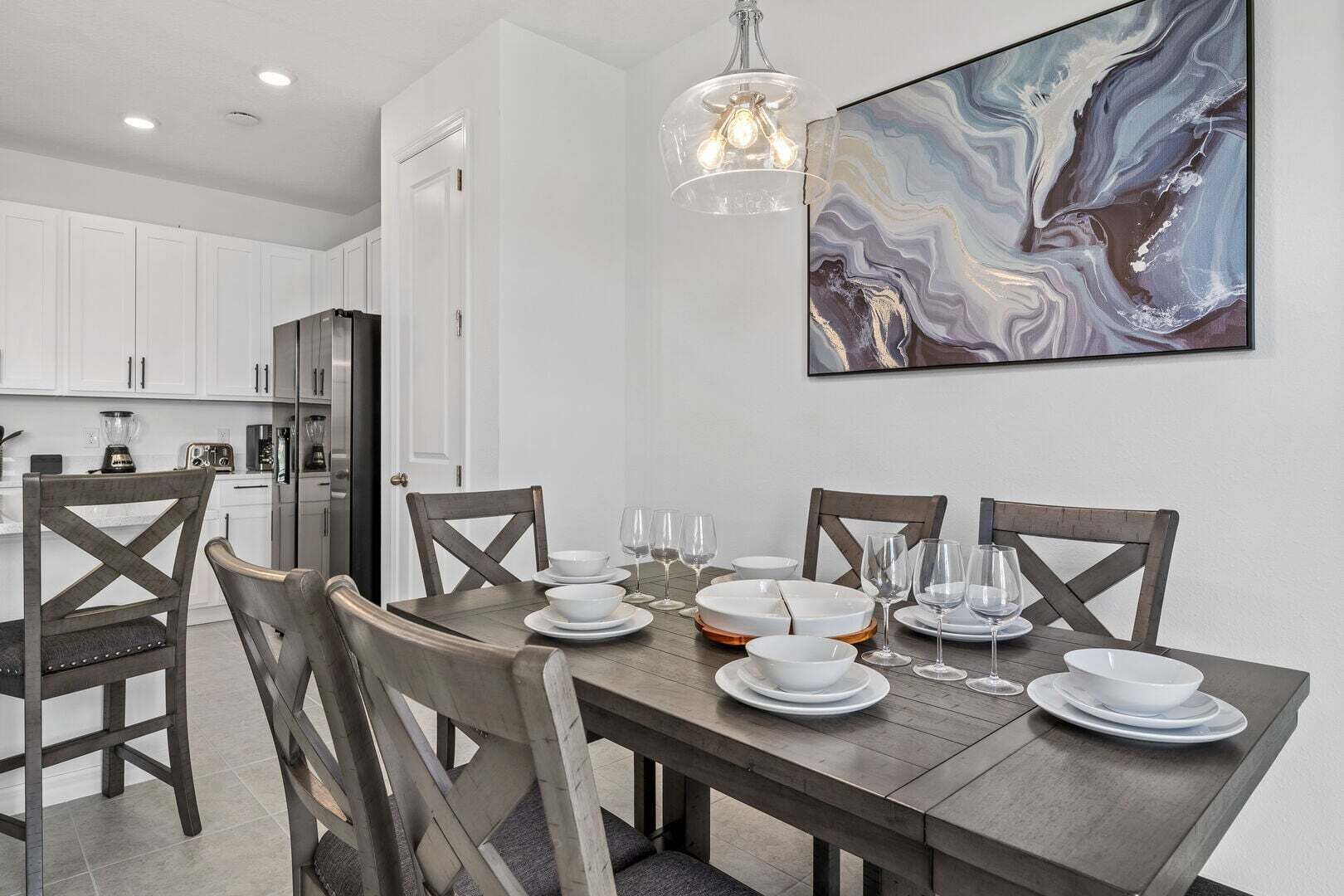 A dining area with a wooden table set for four, white plates, and glasses. There is a modern kitchen in the background with white cabinets and a refrigerator.
