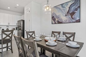 Een eetgedeelte met een houten tafel voor vier personen, witte borden en glazen. Achtergrond toont een moderne keuken met witte kasten en een koelkast.