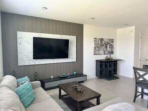 Een woonkamer met een grote flatscreen televisie bevestigd aan een wand met een marmerachtige textuur, een donker houten salontafel, een grijze bank met turquoise kussens, en een grijs kastje met decoratieve objecten.