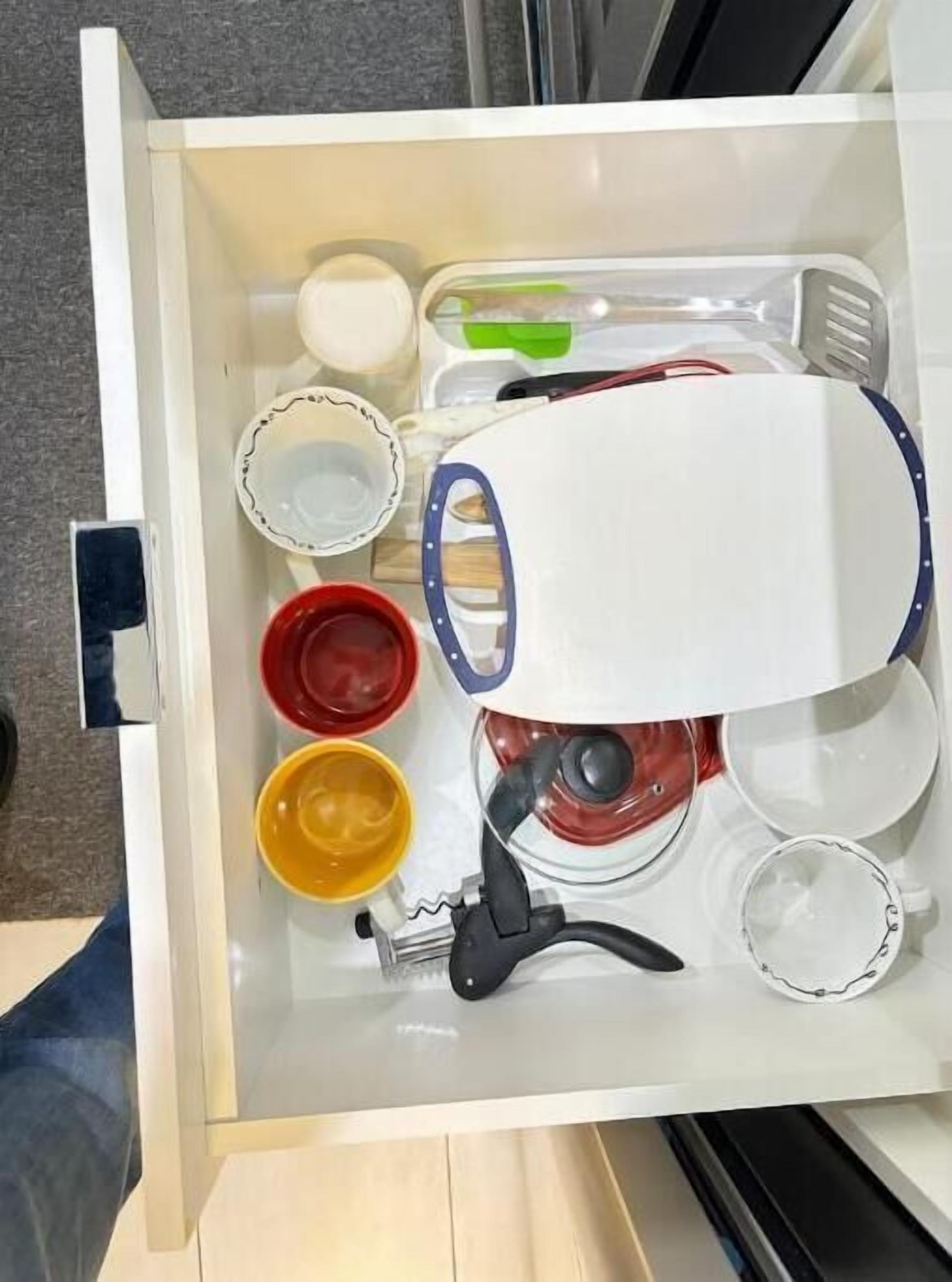 A drawer with various kitchen utensils and containers, including a white pot, red and yellow bowls, and a blue-handled utensil.