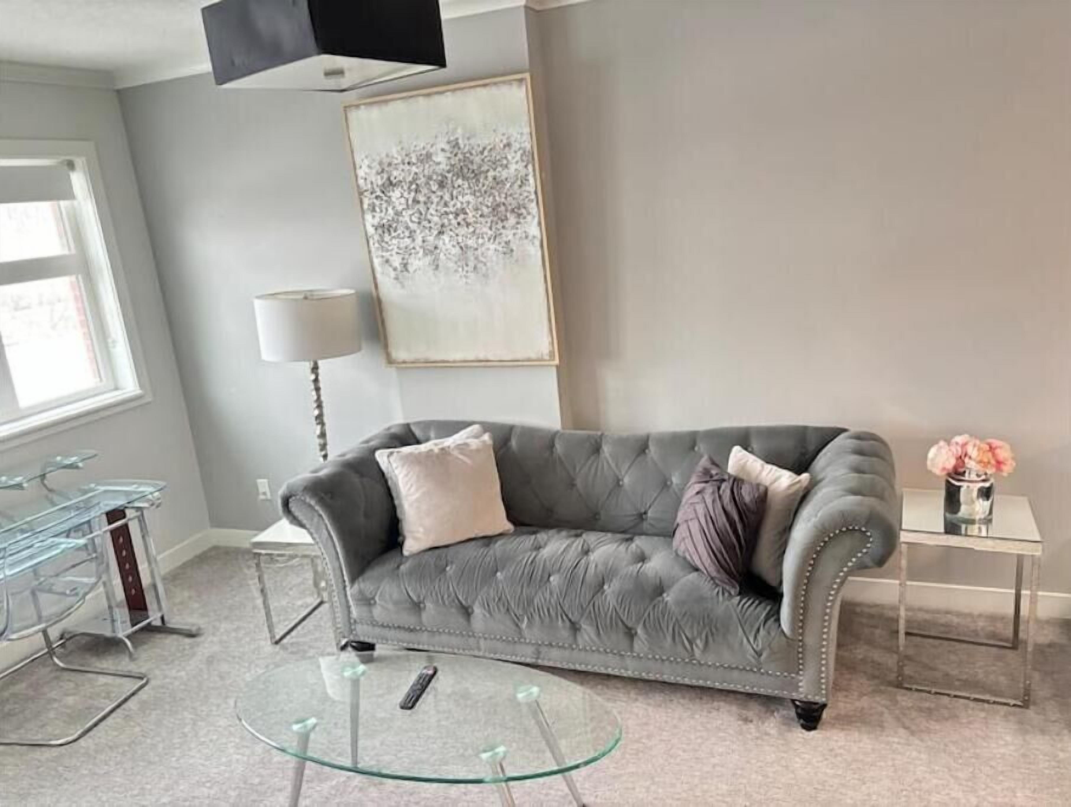Een moderne woonkamer met een grijze gestofte bank, een glazen salontafel, een vloerlamp en een lijst met een abstract schilderij.