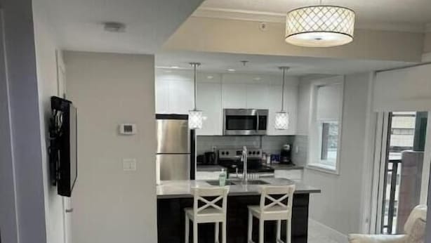 A modern kitchen with white cabinets, stainless steel appliances, and pendant lighting. There is a dining area with a glass table and chairs, and a living room with a sofa and a TV mounted on the wall.