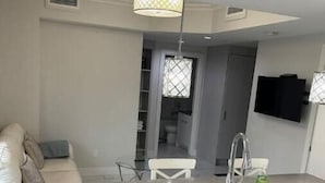 A modern kitchen with a white countertop, stainless steel sink, and a pendant light. There is a white sofa with cushions in the adjacent living area.
