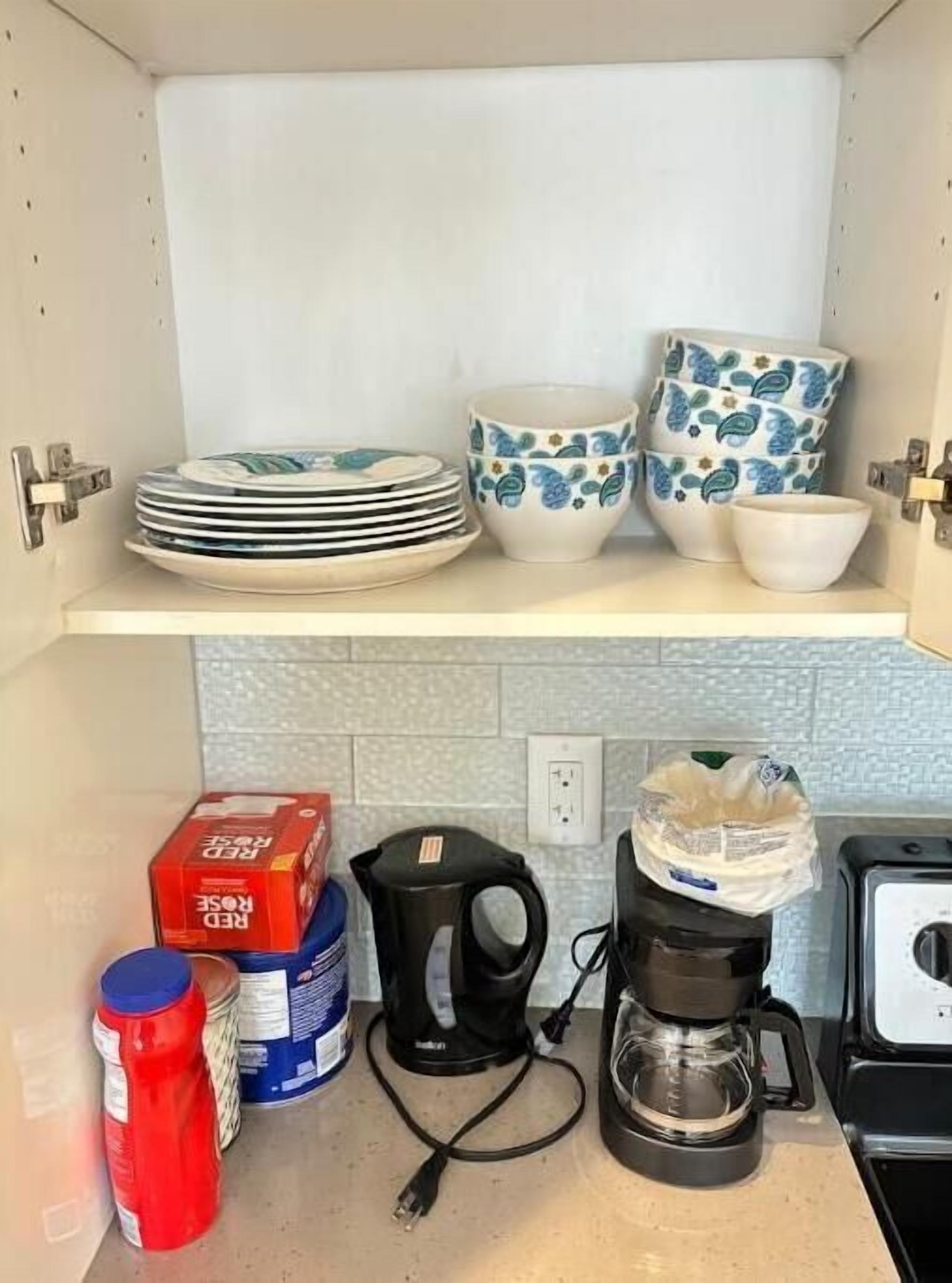 Una cocina con un estante blanco que contiene platos, tazones y una cafetera. Abajo, hay recipientes, una botella roja y un hervidor negro.