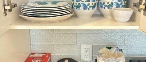 Una cocina con un estante blanco que contiene platos, tazones y una cafetera. Abajo, hay recipientes, una botella roja y un hervidor negro.