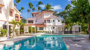 A pool area with a clear blue pool, surrounded by white buildings with red-tiled roofs, and palm trees in the background. - Comfortable & Safe Bavaro Beach Studios (Punta Cana)