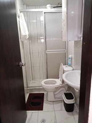 A compact bathroom with a toilet, sink, and shower area, featuring a towel and trash bin.