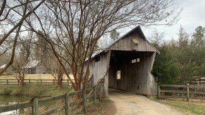 A rustic wooden covered bridge with a dirt path leading to it, surrounded by trees and a wooden fence. - Got Cabin Fever? - Grumpy Ol' Goat (Pleasant Garden)