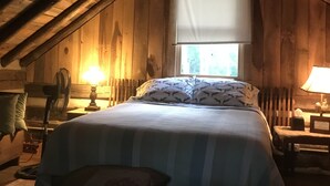 A cozy attic bedroom with a bed, a window with a white blind, wooden beams, a fan, and a lamp. - Got Cabin Fever? - Grumpy Ol' Goat (Pleasant Garden)
