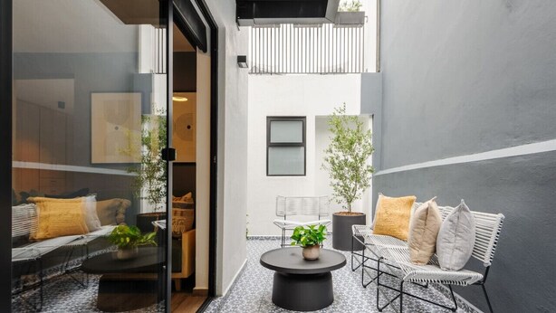Un'area relax esterna moderna con un tavolo rotondo, un divano e sedie, circondata da un pavimento decorato e un muro grigio.