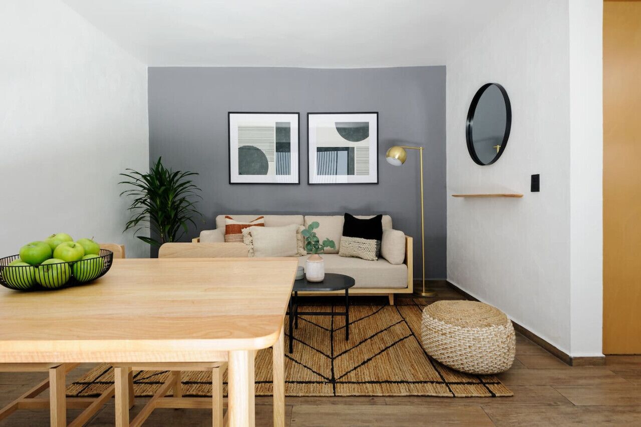 A modern living room with a wooden dining table, a sofa, and a coffee table. There are two framed abstract artworks on the wall, a floor lamp, and a round mirror.