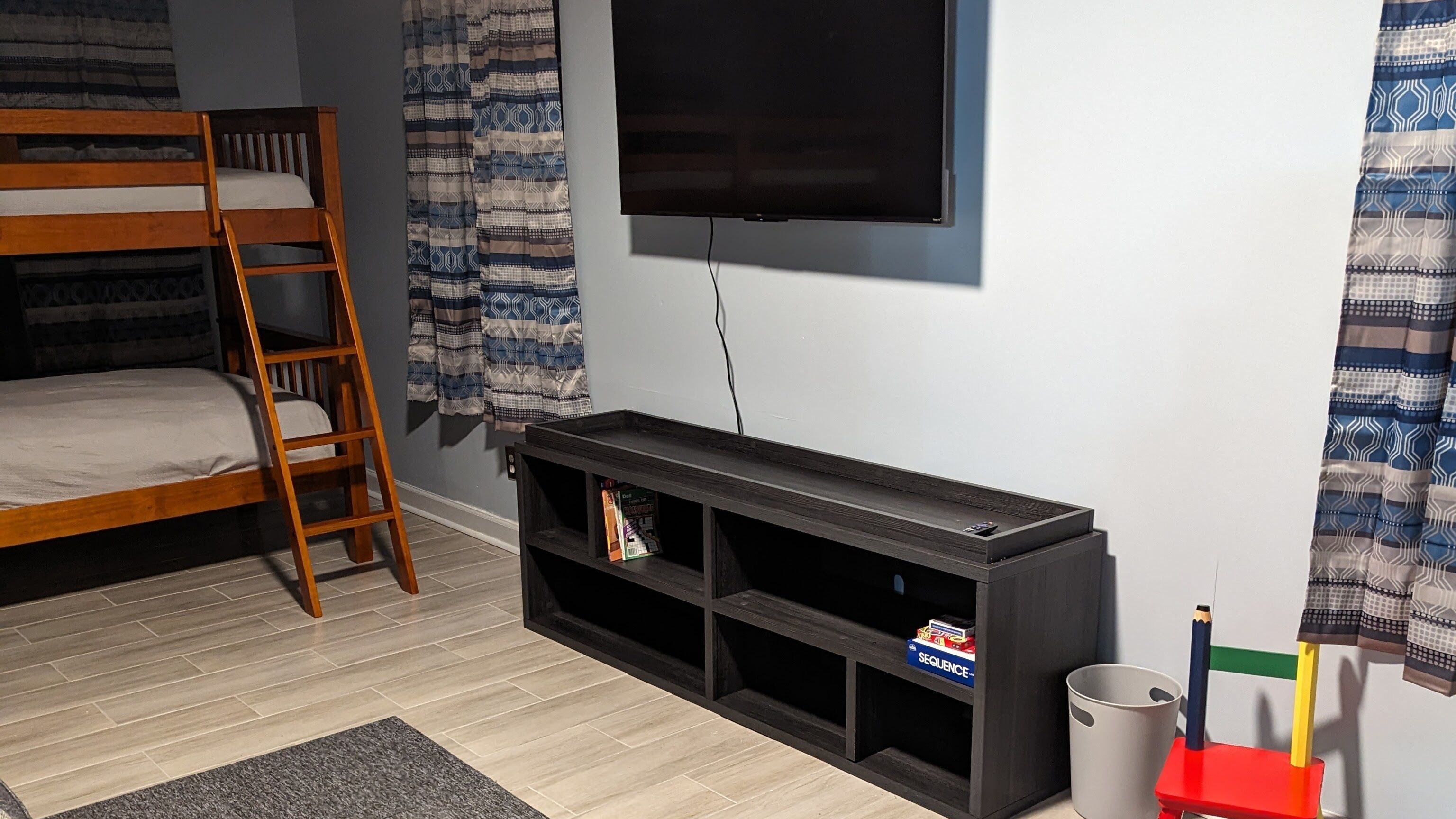 A room with a bunk bed, a TV mounted on the wall, a black entertainment unit, and a small trash can.