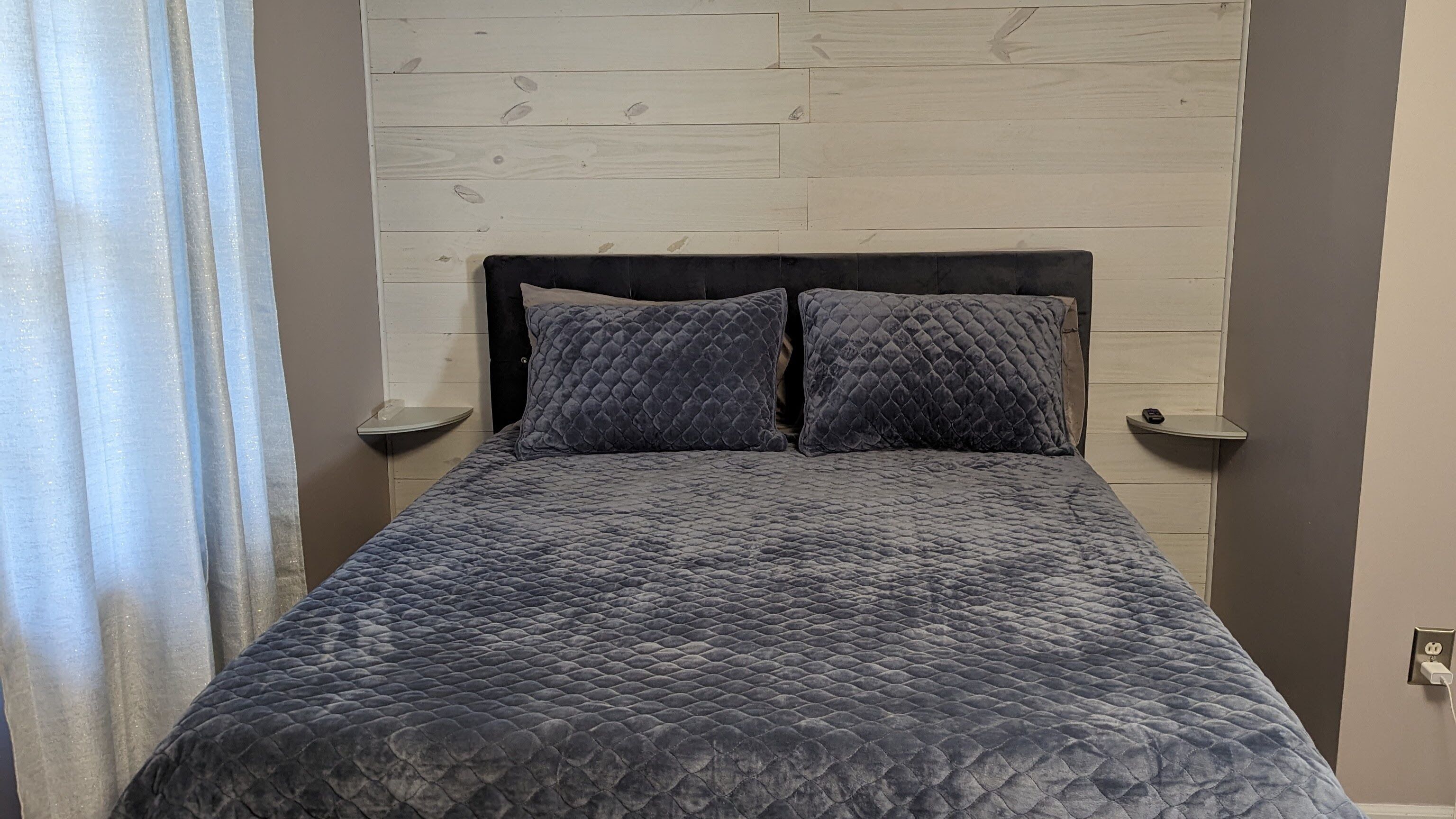 Ein Bett mit einer gesteppten, grauen Bettdecke und zwei Kissen, einem Holz-Kopfteil, Nachttischen und einer Steckdose an der Wand.