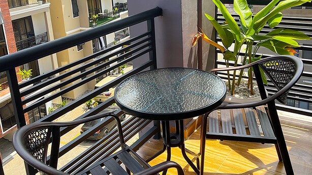 Un balcón con una mesa redonda de vidrio y dos sillas, una planta en maceta y vistas de edificios cercanos.