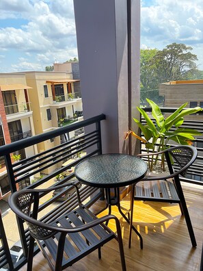A balcony with a round glass table and two chairs, a potted plant, and a view of neighboring buildings.