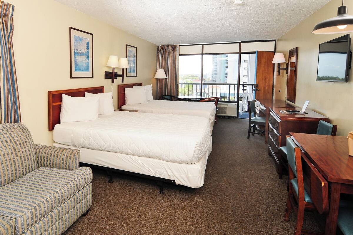 A hotel room with two beds, a sofa, a desk, and a balcony with a view of buildings.