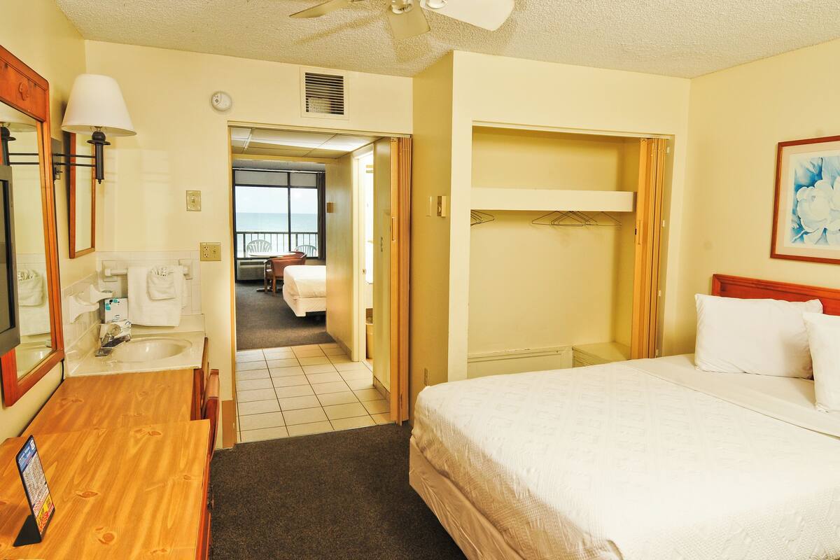 A hotel room with a bed, a desk with a lamp, a bathroom with a sink, and a view of the ocean through an open door.