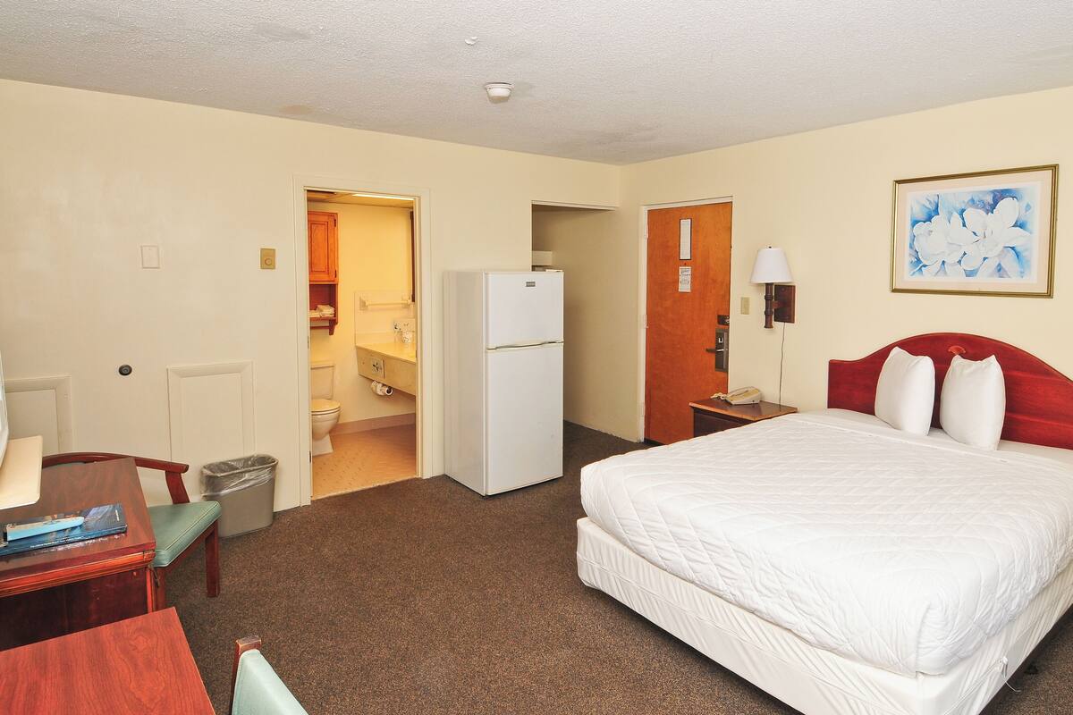 A hotel room with a bed, a desk, a chair, a refrigerator, a bathroom, and a painting on the wall.