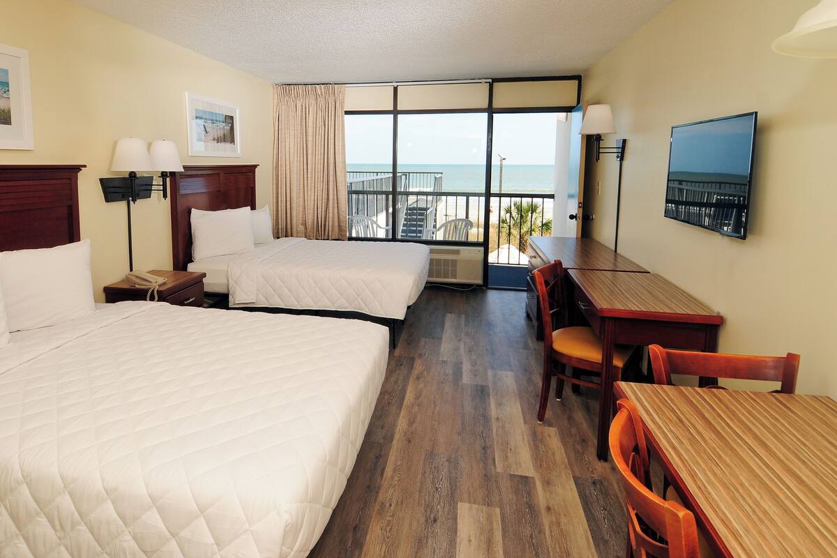 A hotel room with two beds, a desk, a chair, a TV, and a balcony with a view of the sea.