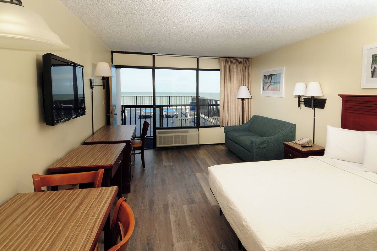 A hotel room with a bed, a desk with a chair, a green armchair, a TV, and a window with a view of the beach.