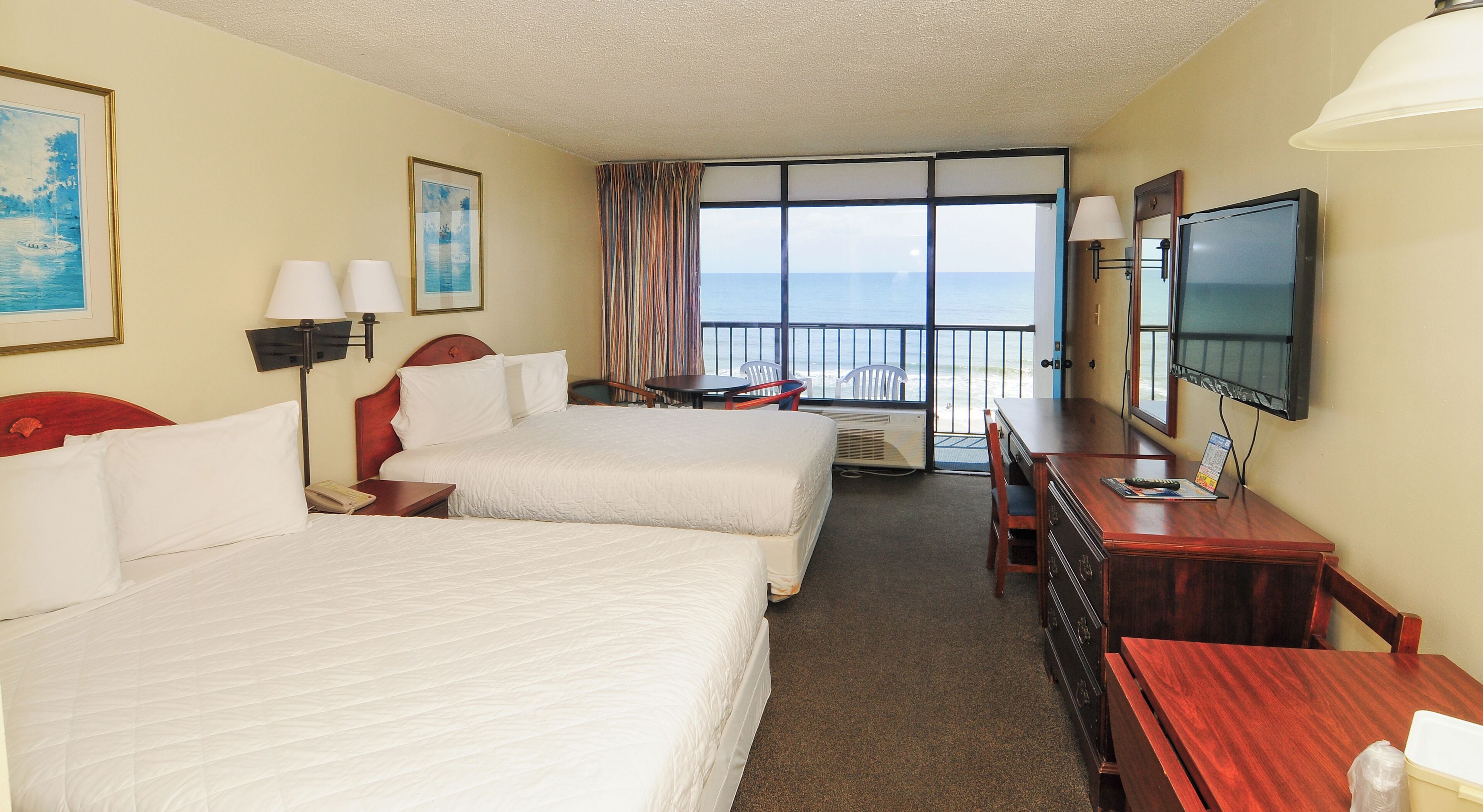 A hotel room with two beds, a TV, a desk, and a balcony with a view of the sea.