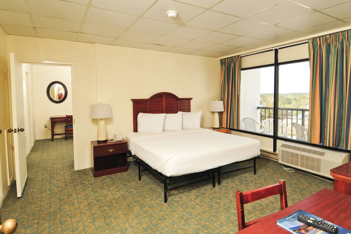 A hotel room with a large bed, bedside lamps, a nightstand, a desk with a chair, a window with curtains, and a view of the outdoors.