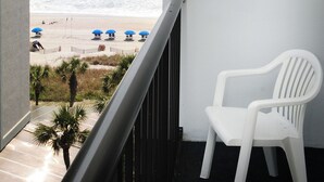Un balcón con una silla de plástico blanca, una pared blanca y vistas a la playa con sombrillas azules.