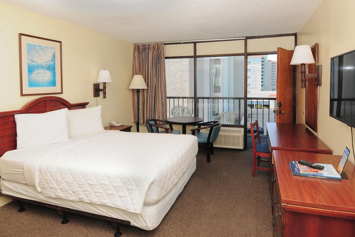 A hotel room with a bed, a desk, a chair, a table, a TV, and a window with a view of buildings.