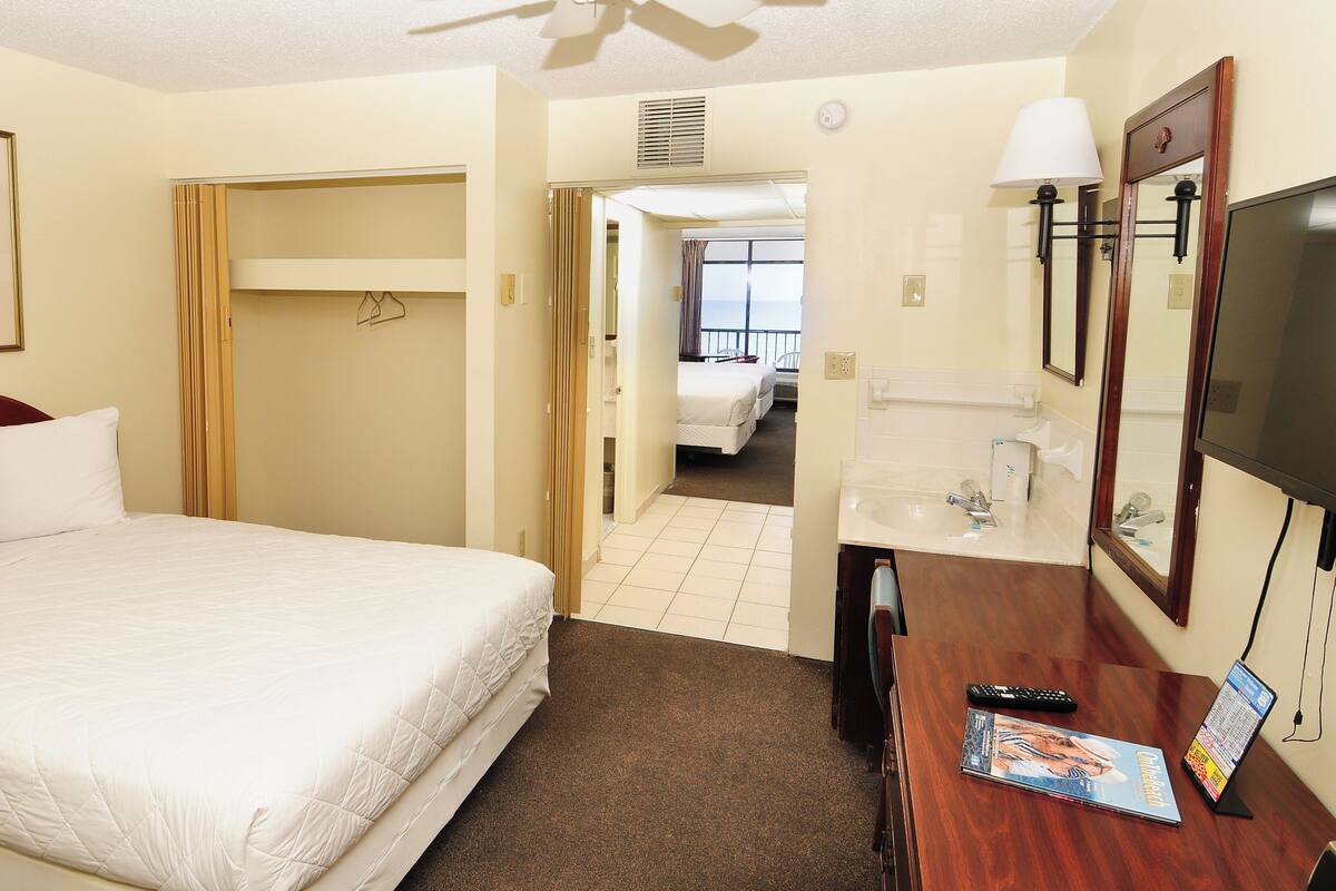A hotel room with a bed, a desk with a computer and remote, a TV, a bathroom with a mirror, and a painting on the wall.