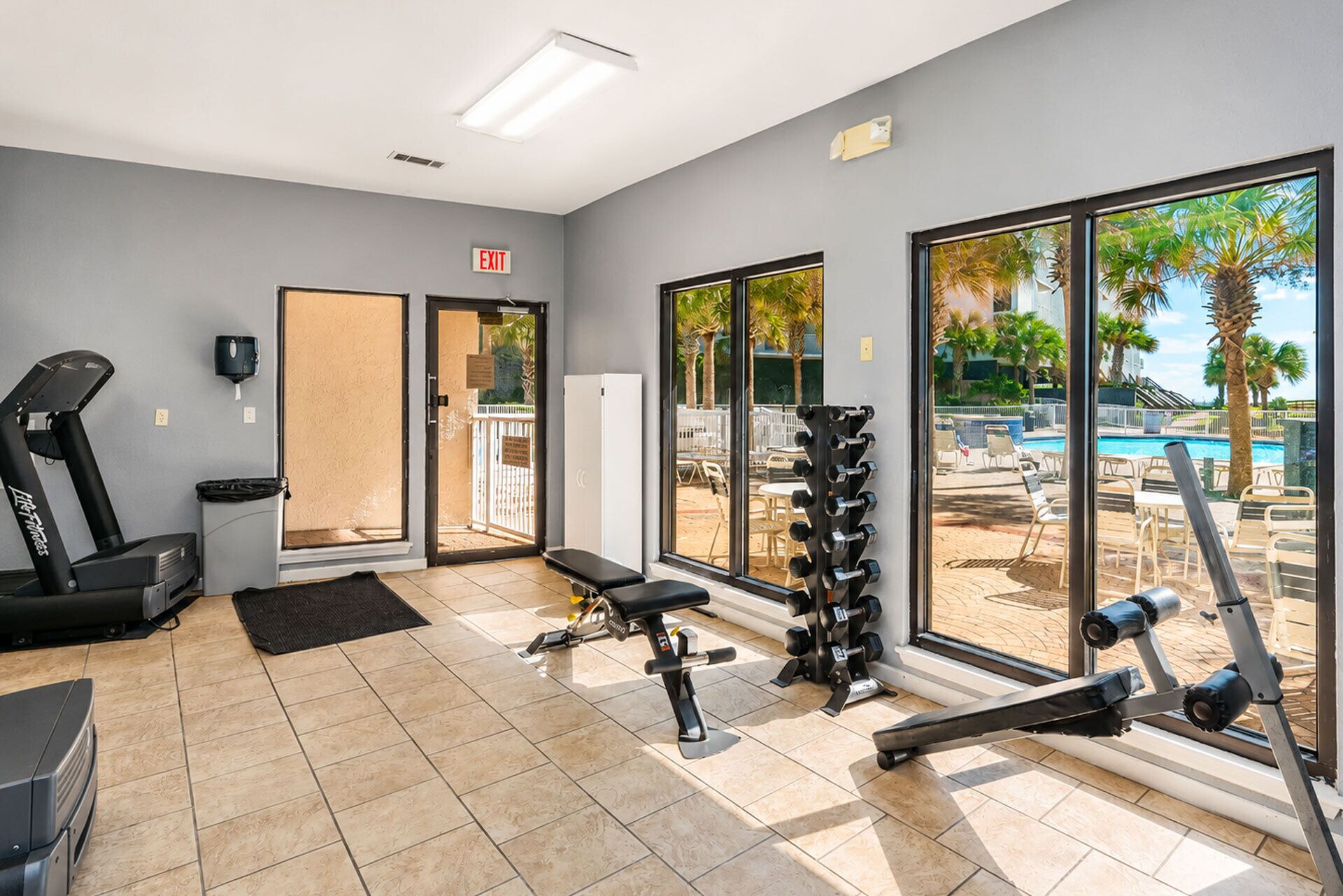 A gym with exercise equipment, a treadmill, and a view of a pool and palm trees.