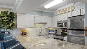 Una cocina con armarios blancos, encimera de mármol, refrigerador de acero inoxidable y un letrero que dice "BEACH HOUSE".
