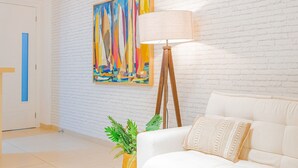 Una habitación con pared de ladrillo blanco, un cuadro abstracto colorido, una lámpara de pie, un sofá blanco y una planta en maceta.
