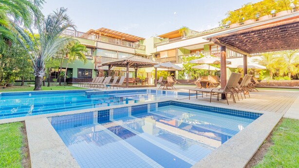 Una piscina con sillones de descanso y sombrillas, rodeada de vegetación y un edificio de varios pisos.