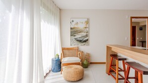Une pièce comprenant une table à manger en bois, une chaise en osier, un pouf en osier et un tableau représentant une scène de plage, accroché au mur.
