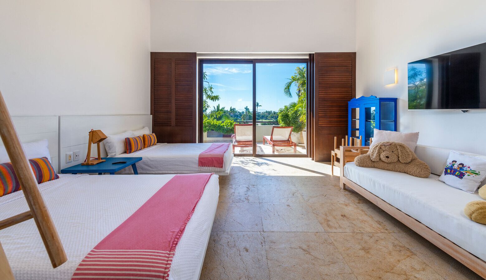 A hotel room with two beds, a sofa, a TV, and a balcony with a view of greenery.