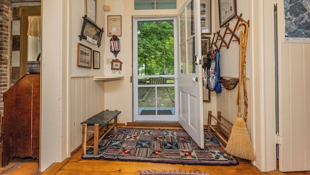 A hallway with a wooden bench, a patterned rug, a broom leaning against the wall, and a door leading outside. - Historic Parker’s Landing
(Springfield)