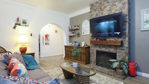 A cozy living room with a fireplace, a flat-screen TV mounted above it, a comfortable sofa with colorful cushions, and a coffee table. - Versatile Downtown 1B1B Home near Disneyland (Santa Ana)