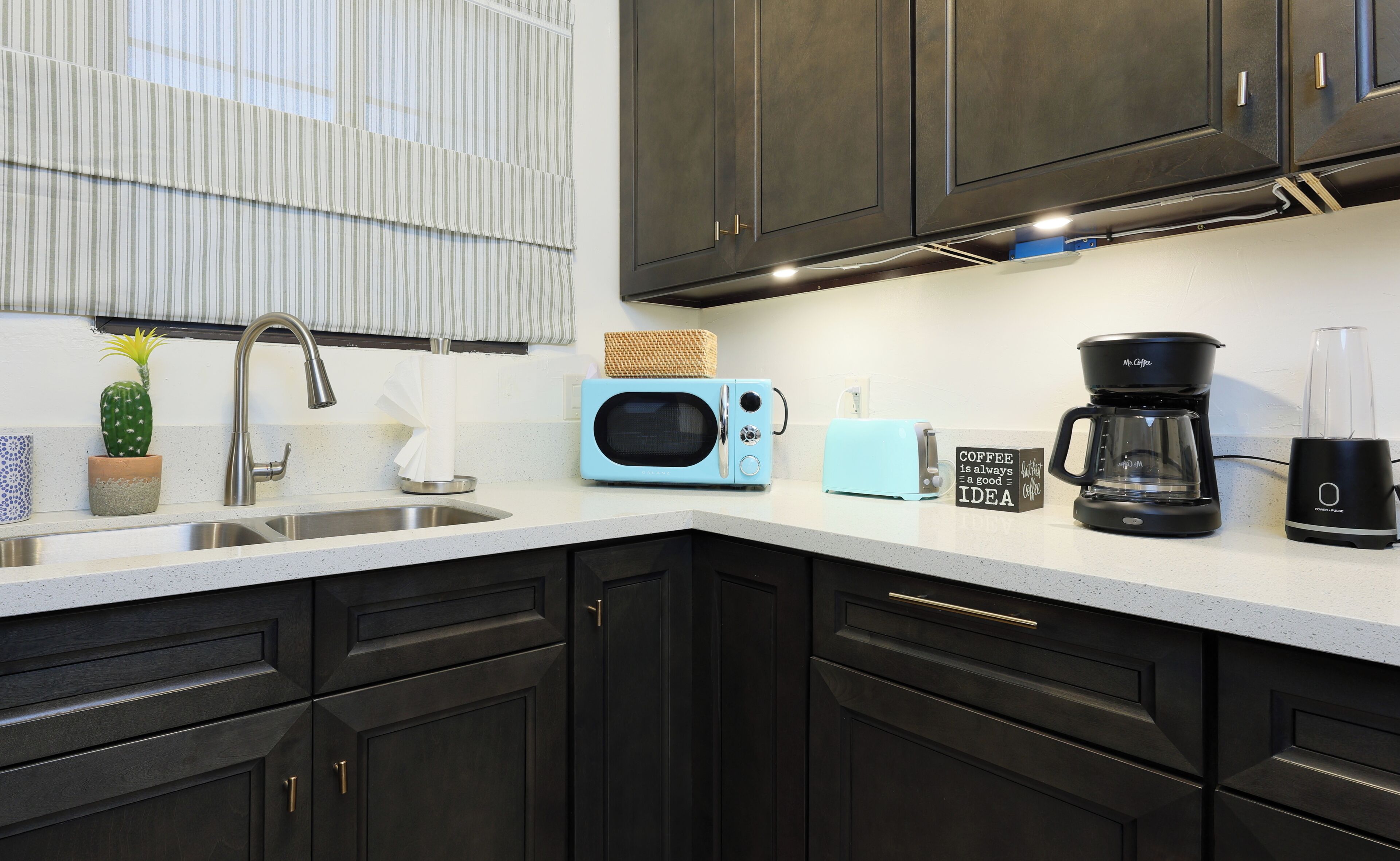 A modern kitchen with dark cabinets, a stainless steel sink, a blue microwave, a coffee maker, and a toaster.