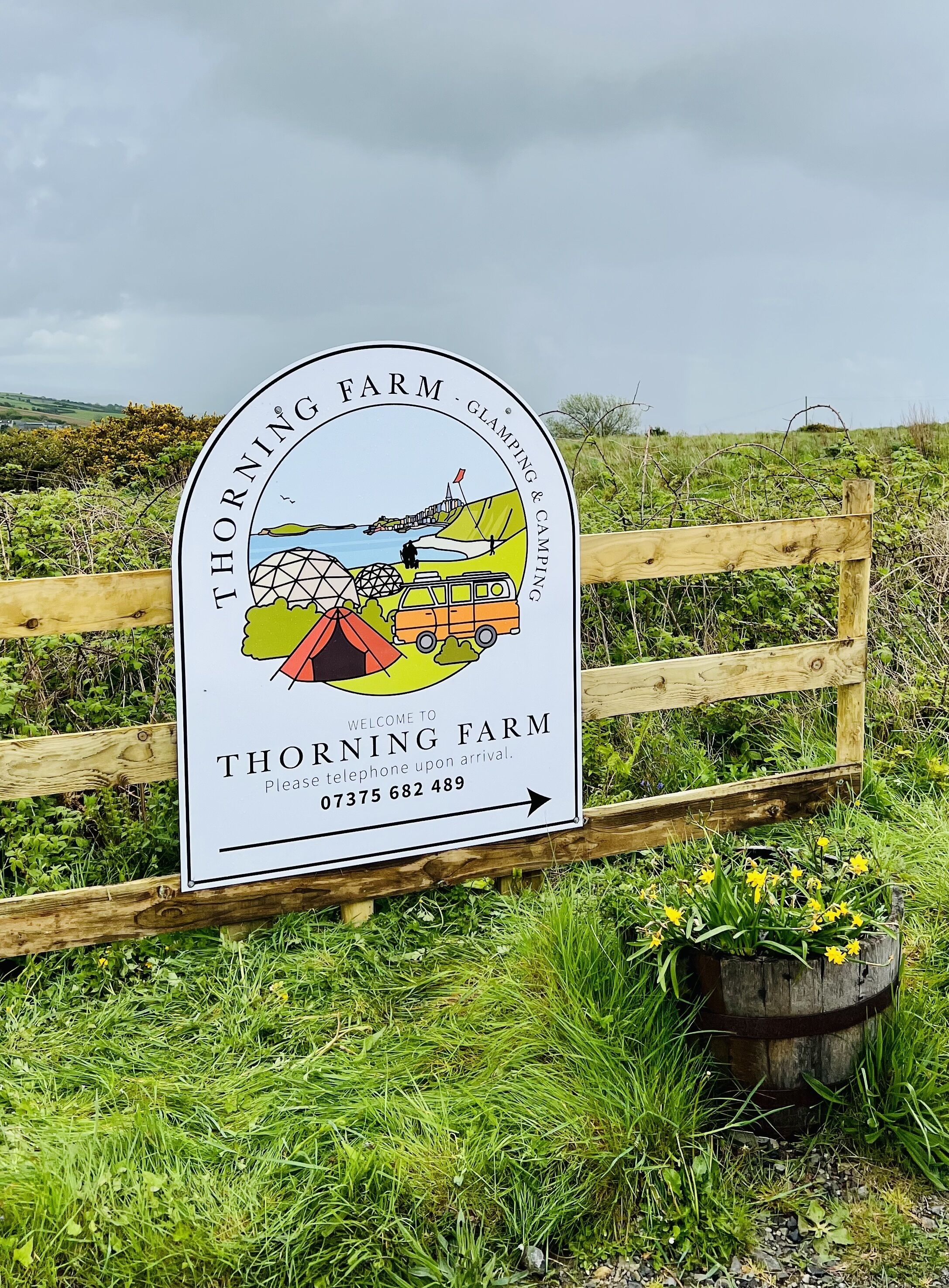 A welcome sign for Thorning Farm, featuring a camping scene with a tent, van, and person fishing.