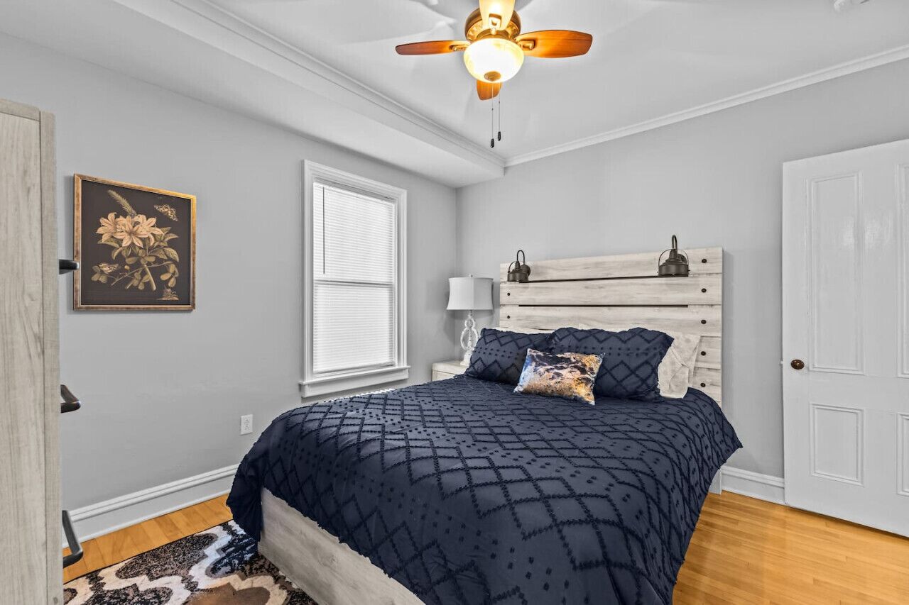 A bedroom with a bed, a ceiling fan, a window with blinds, a wall-mounted lamp, and a door.