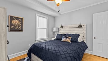 A bedroom with a bed, a ceiling fan, a window with blinds, a wall-mounted lamp, and a door.