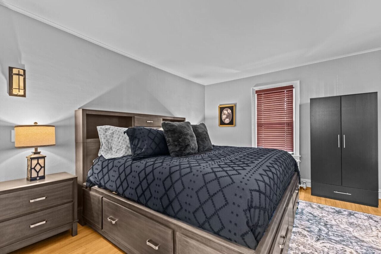 A bedroom with a bed, a nightstand, a lamp, a wardrobe, and a framed picture on the wall.