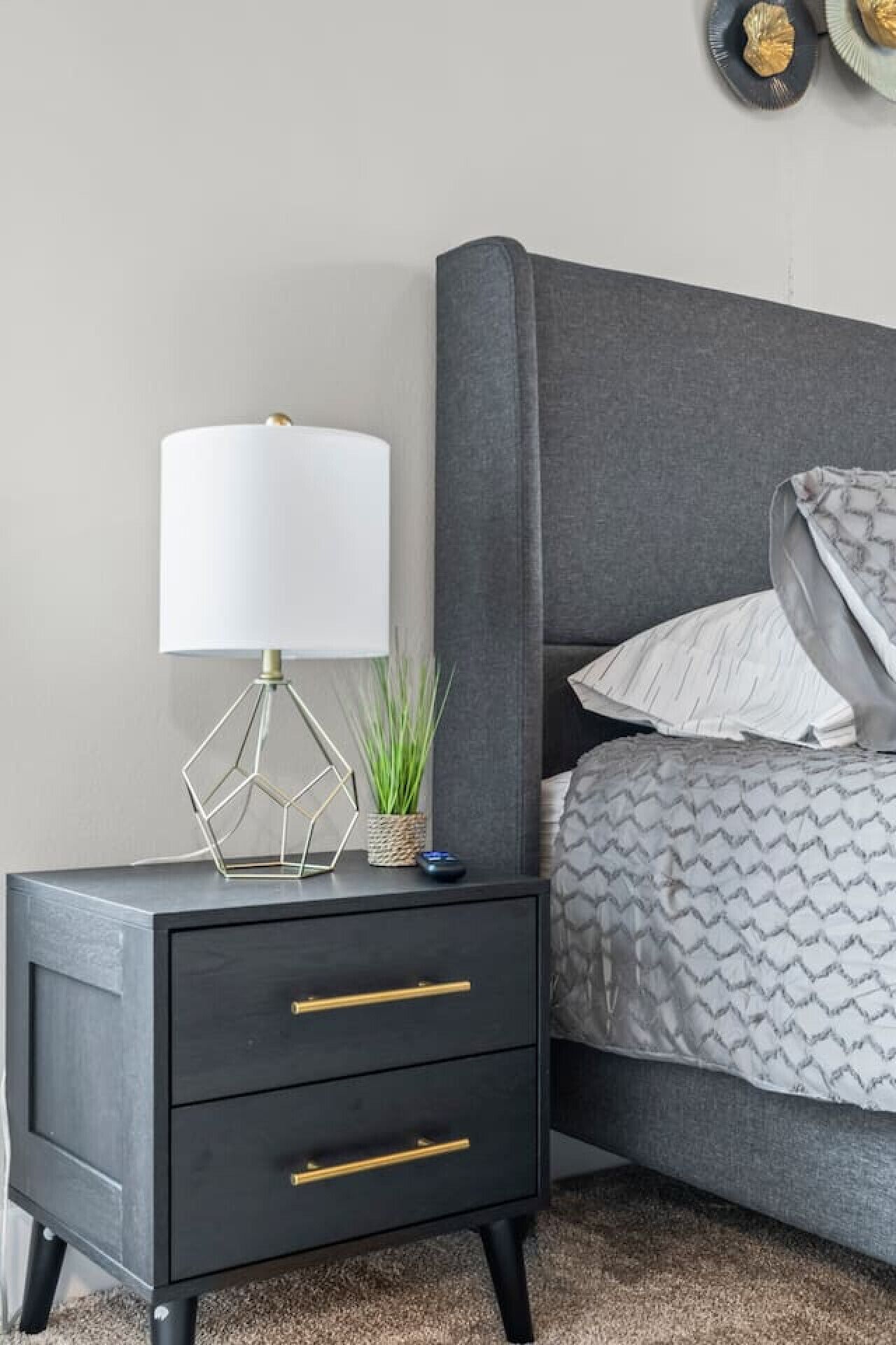 A modern bedroom with a grey upholstered bed, a bedside table with a geometric lamp, and a small potted plant.