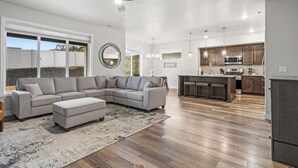 Un salon moderne avec un canapé d’angle, un coin repas doté de tabourets de bar et une cuisine avec des meubles en bois et des appareils électroménagers en acier inoxydable.