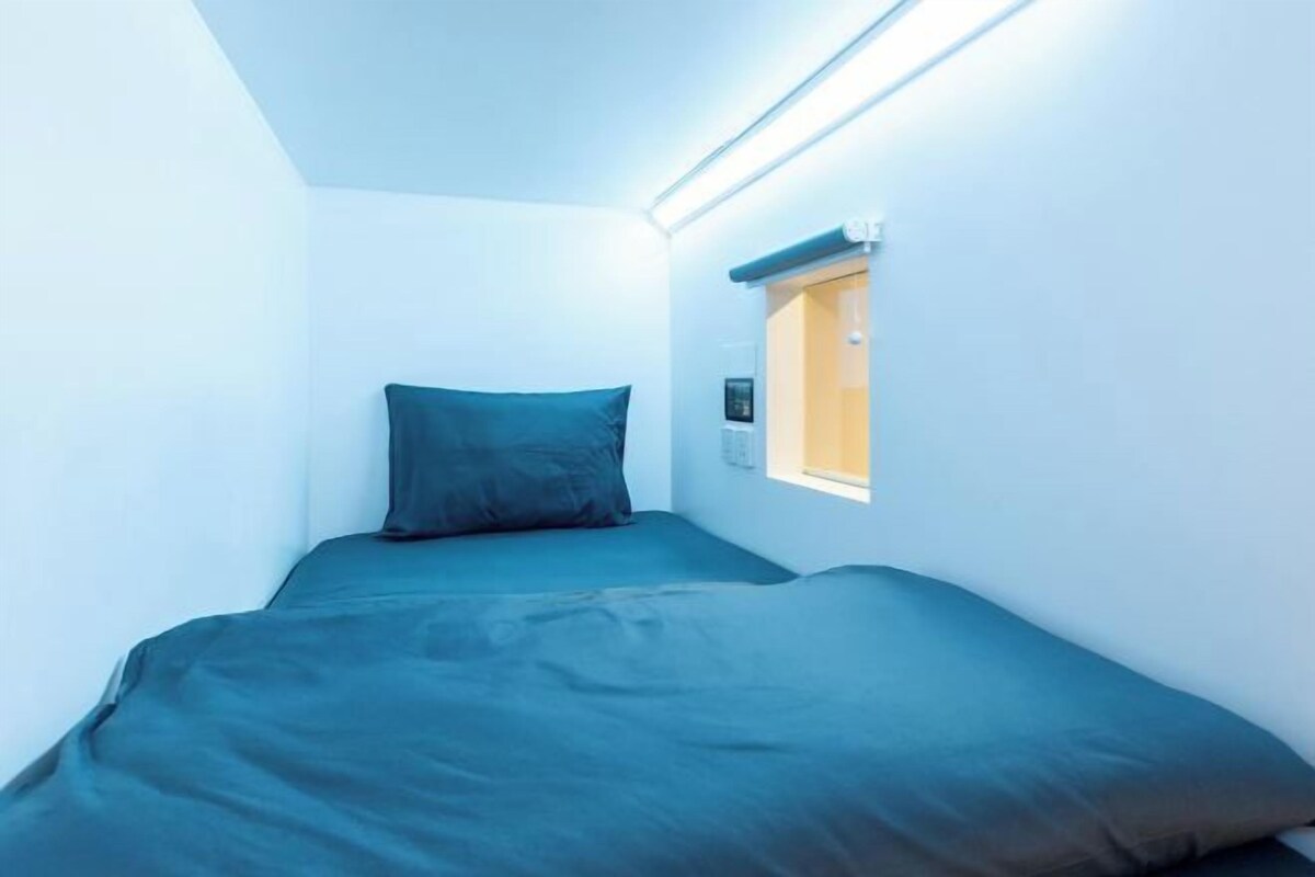 A small, narrow room with a single bed, a window with a view, and a wall-mounted light fixture.