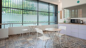 Cocina moderna con microondas, hervidor y lavabo. Hay sillas blancas y una mesa frente a ventanas amplias con vista a árboles.