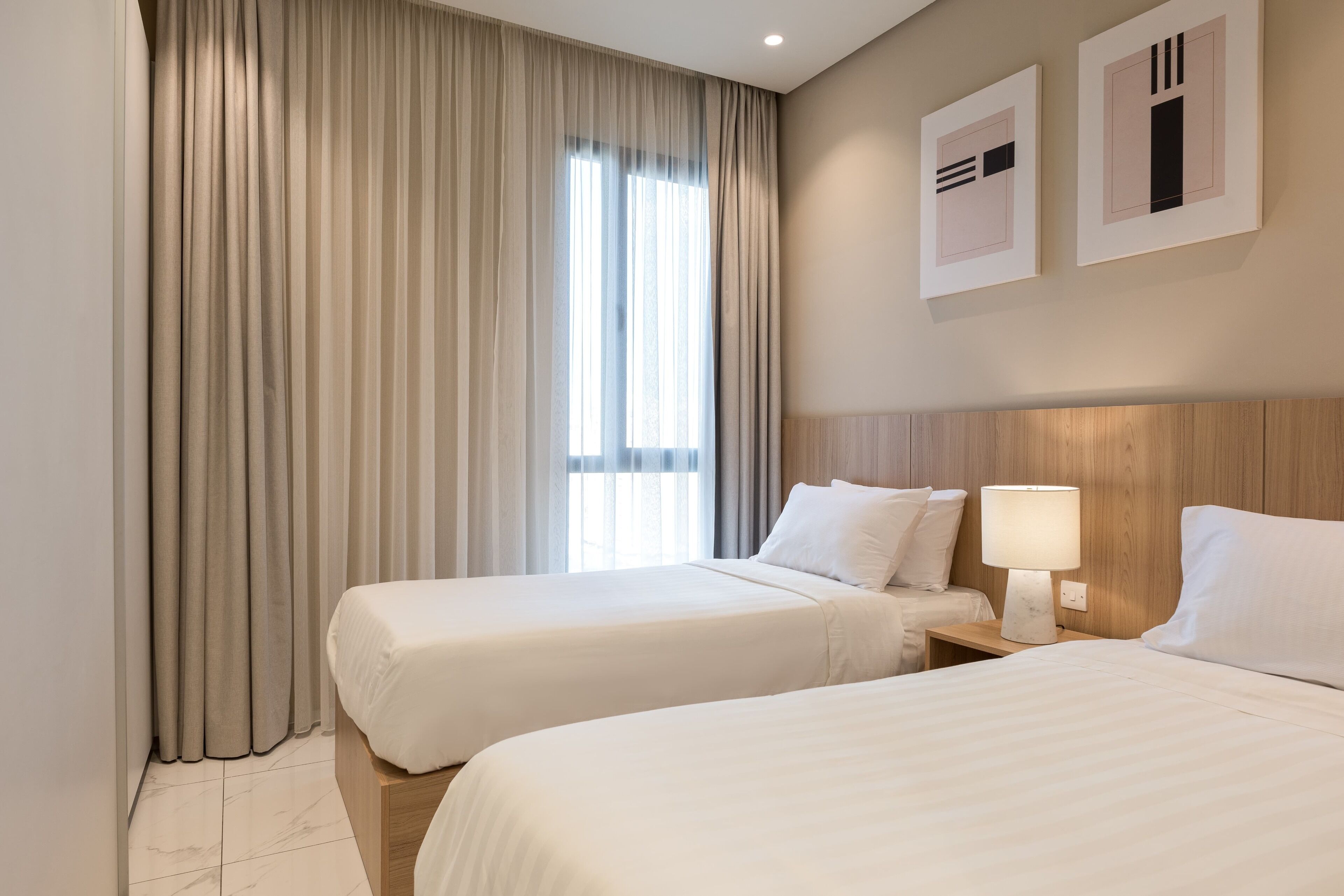 A hotel room with two beds, a large window with curtains, a wooden headboard, a nightstand with a lamp, and two framed pictures on the wall.