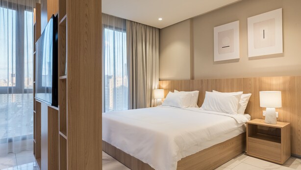 Une chambre d’hôtel moderne dotée d’un grand lit, d’une tête de lit en bois et offrant une vue sur la ville grâce à de grandes fenêtres du sol au plafond.